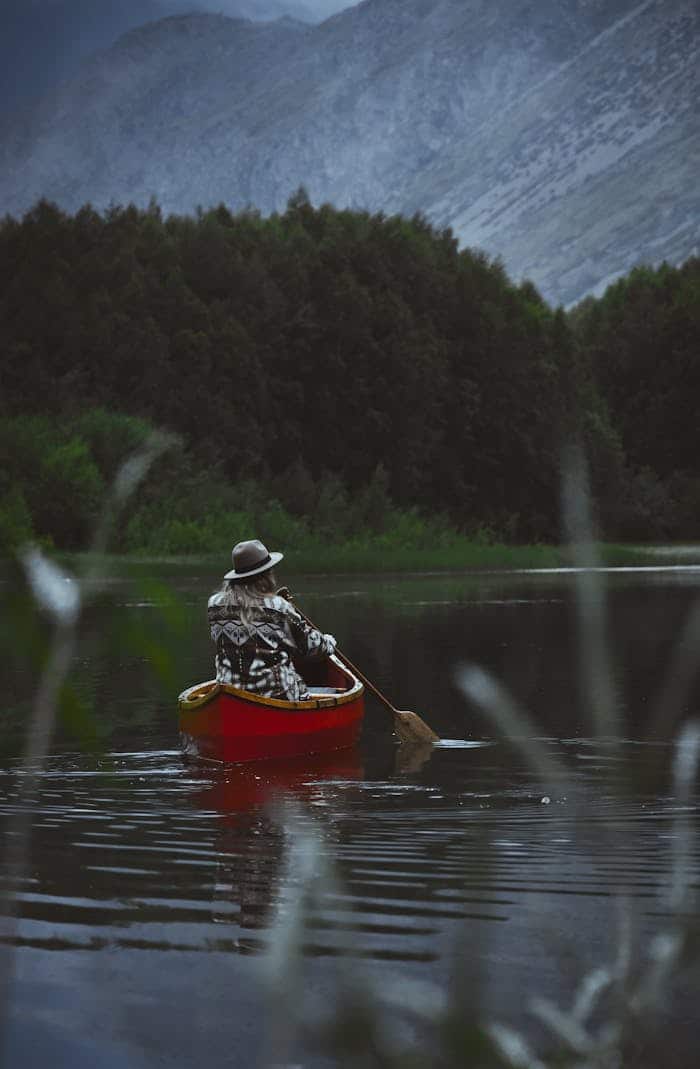 mission-02 Person canoeing in a serene mountain lake surrounded by lush forest and towering peaks.